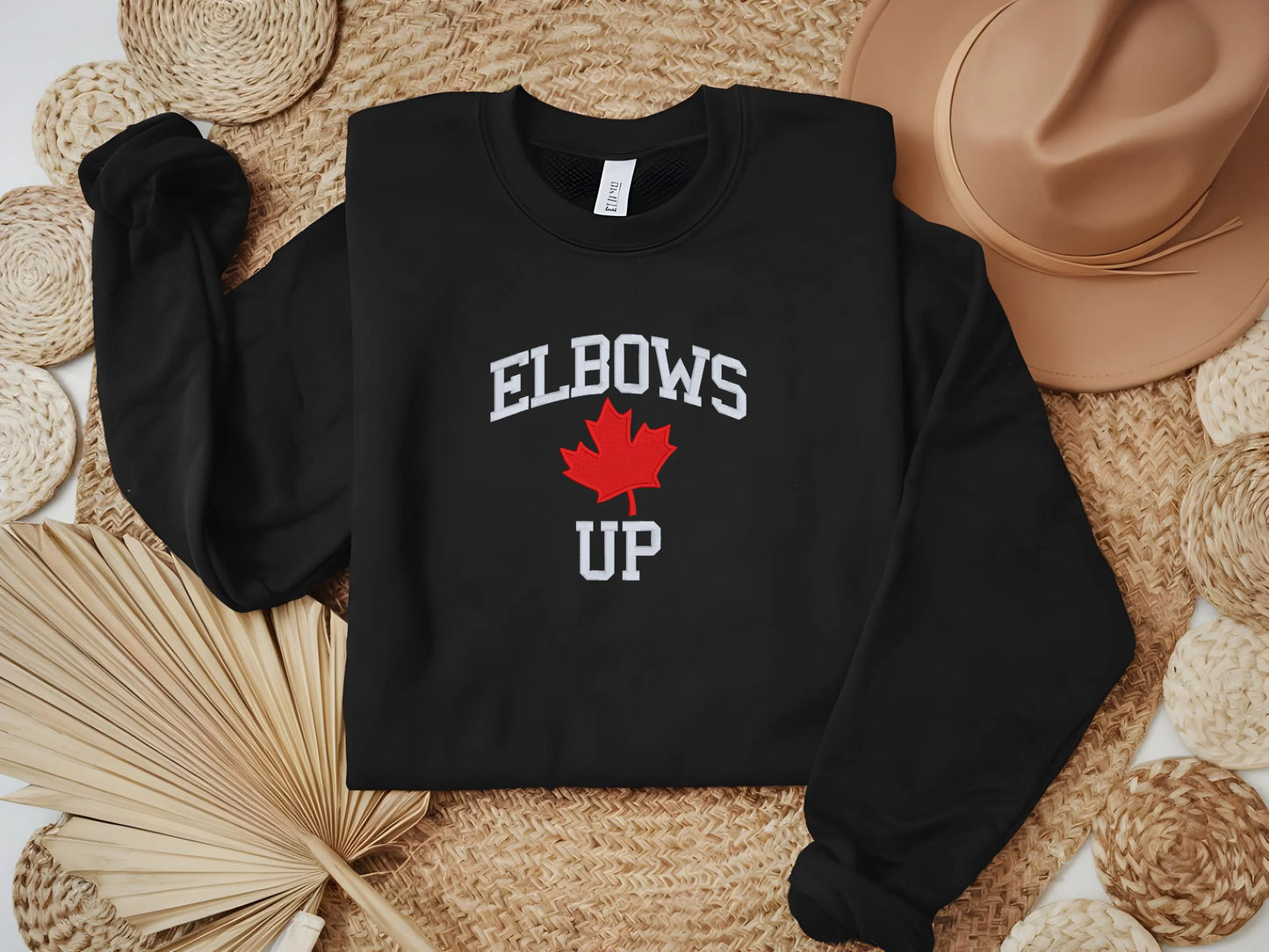 Black sweatshirt with 'ELBOWS UP' and a maple leaf design on a woven surface.