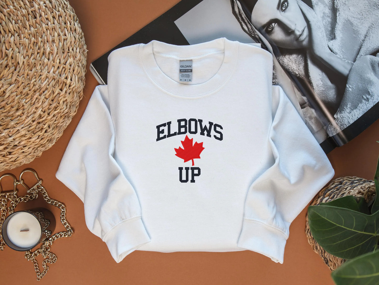 White sweatshirt with 'ELBOWS UP' text and red maple leaf on a brown surface.