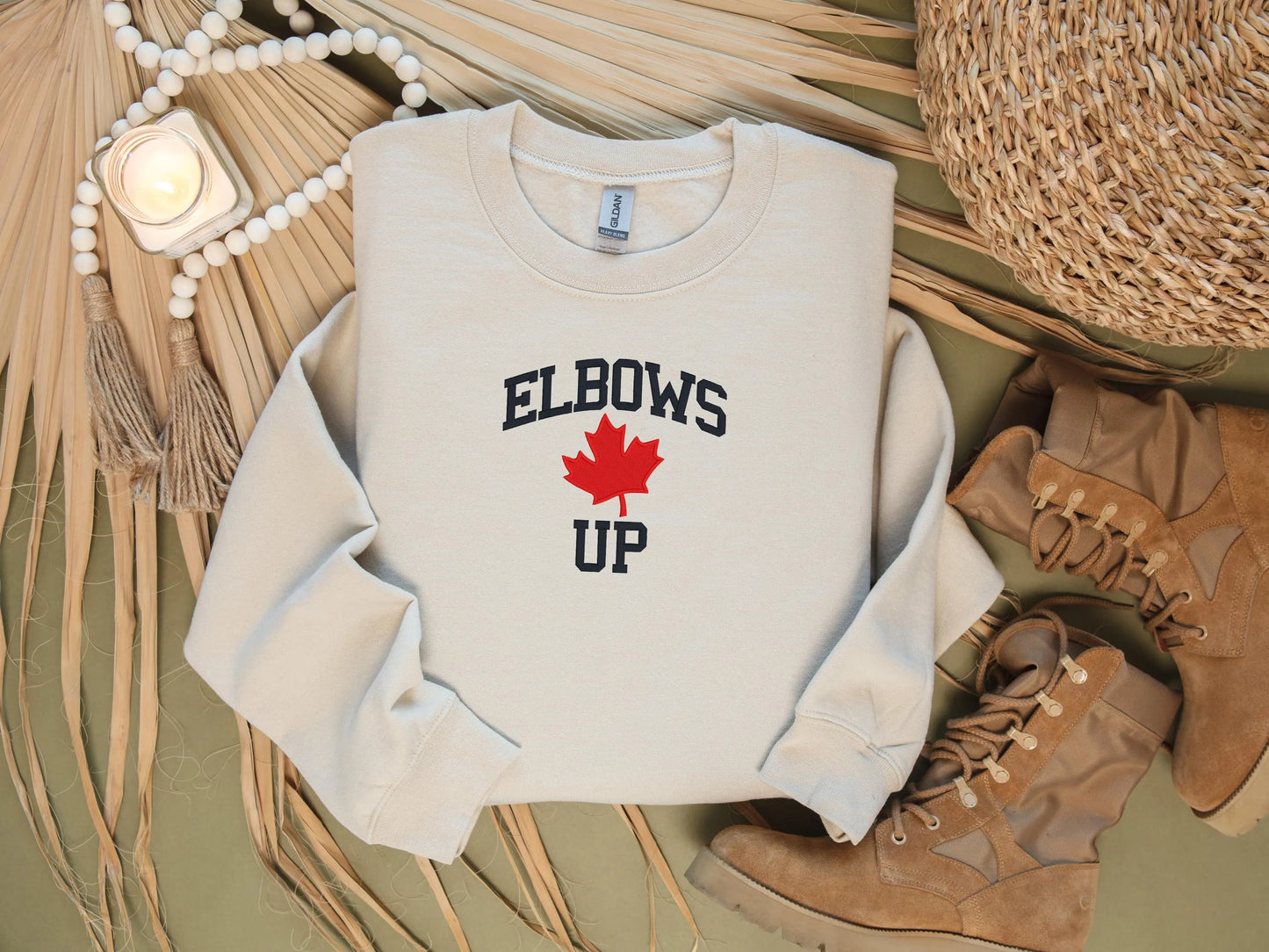 Beige sweatshirt with 'ELBOWS UP' text and red maple leaf graphic on a textured surface with boots and decor items.