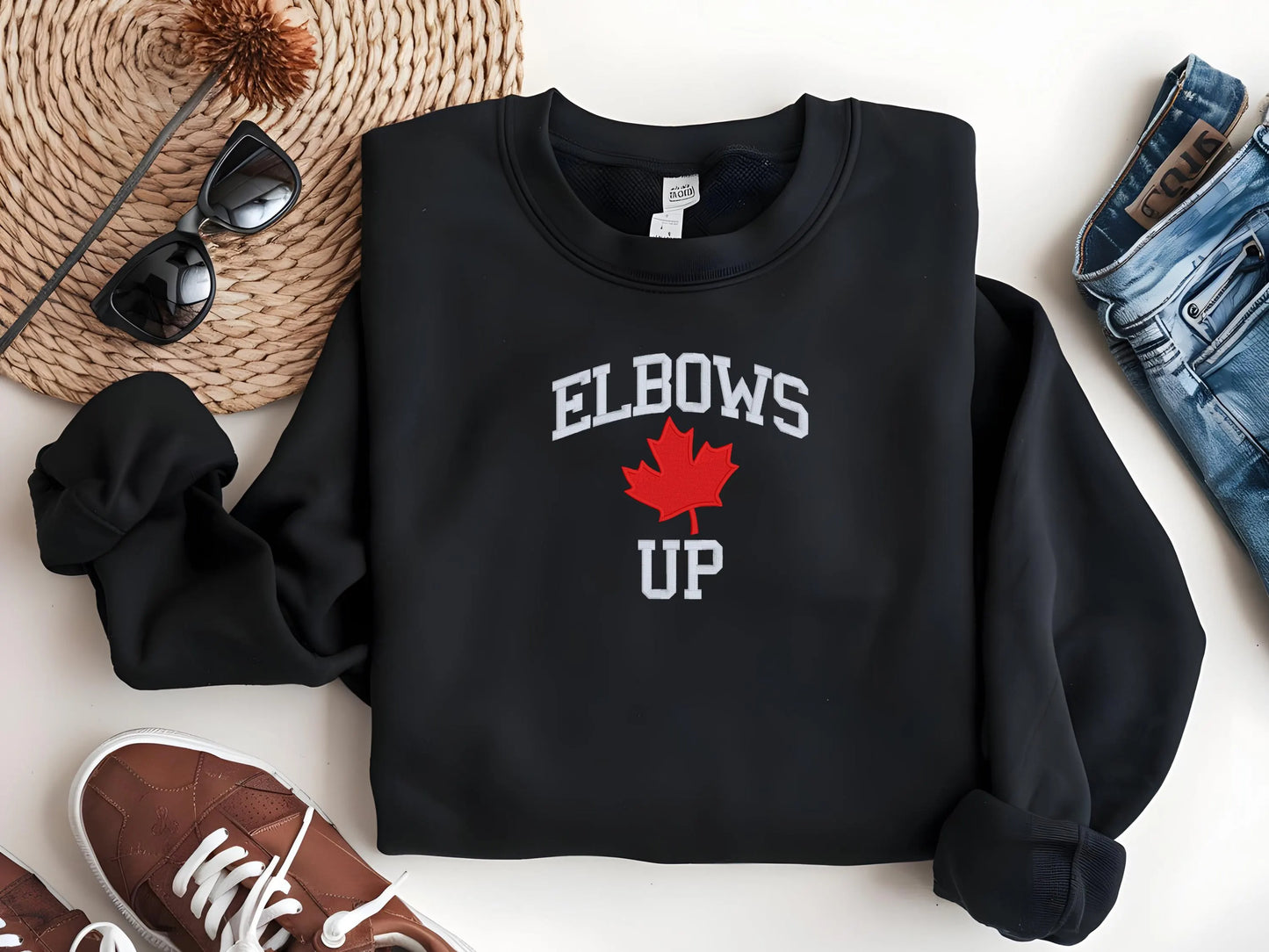 Black sweatshirt with 'ELBOWS UP' and a maple leaf graphic on a white surface with accessories.