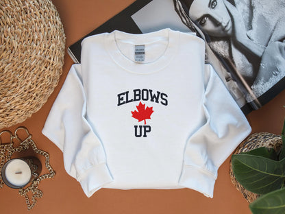 White sweatshirt with 'ELBOWS UP' text and red maple leaf on a brown surface.